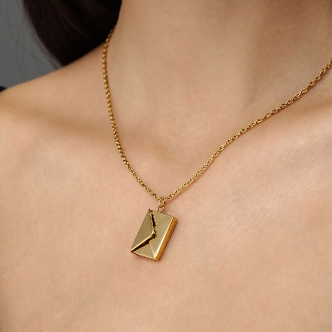 Un primer plano de una persona que lleva el collar "Mi carta de amor", una cadena de oro con un colgante en forma de sobre y una abertura en zigzag, destaca su diseño único, que lo convierte en un atento regalo para esposa.