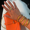Una mano con uñas de color rosa palo lleva anillos de oro, tres brazaletes y el Collar moldeable multiuso de acero inoxidable, descansa sobre tela blanca texturizada mientras sostiene un bolso naranja con una correa de cadena dorada.