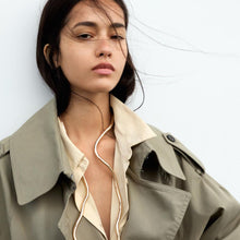 Cargar imagen en el visor de la galería, Una mujer de pelo largo y oscuro lleva una gabardina abierta sobre una camisa beige y un Multiuso Moldeable al Cuello. Mira con confianza a la cámara mientras los mechones sueltos le revuelven la cara, resaltando este versátil accesorio sobre un fondo liso.