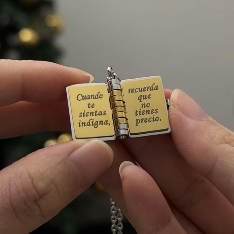 Una persona sostiene el "Collar para hija - Mi Pequeña Estrella", un medallón en forma de libro con texto en español: "Cuando te sientas indigna, recuerda que no tienes precio". Al fondo se ve borroso un árbol de Navidad.