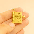 Una mano sostiene el Collar para hija - Mi Pequeña Estrella, un medallón de oro con forma de pequeño libro grabado con "Mi Pequeña Estrella" y un corazón. Este collar de oro incluye una pequeña estrella y una anilla para enganchar a una cadena: un regalo significativo.