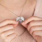 Una persona sostiene un collar personalizado con foto en forma de corazón plateado, abierto para mostrar una pequeña foto de dos personas en su interior. Lleva puesto un top rosa suave y esponjoso.