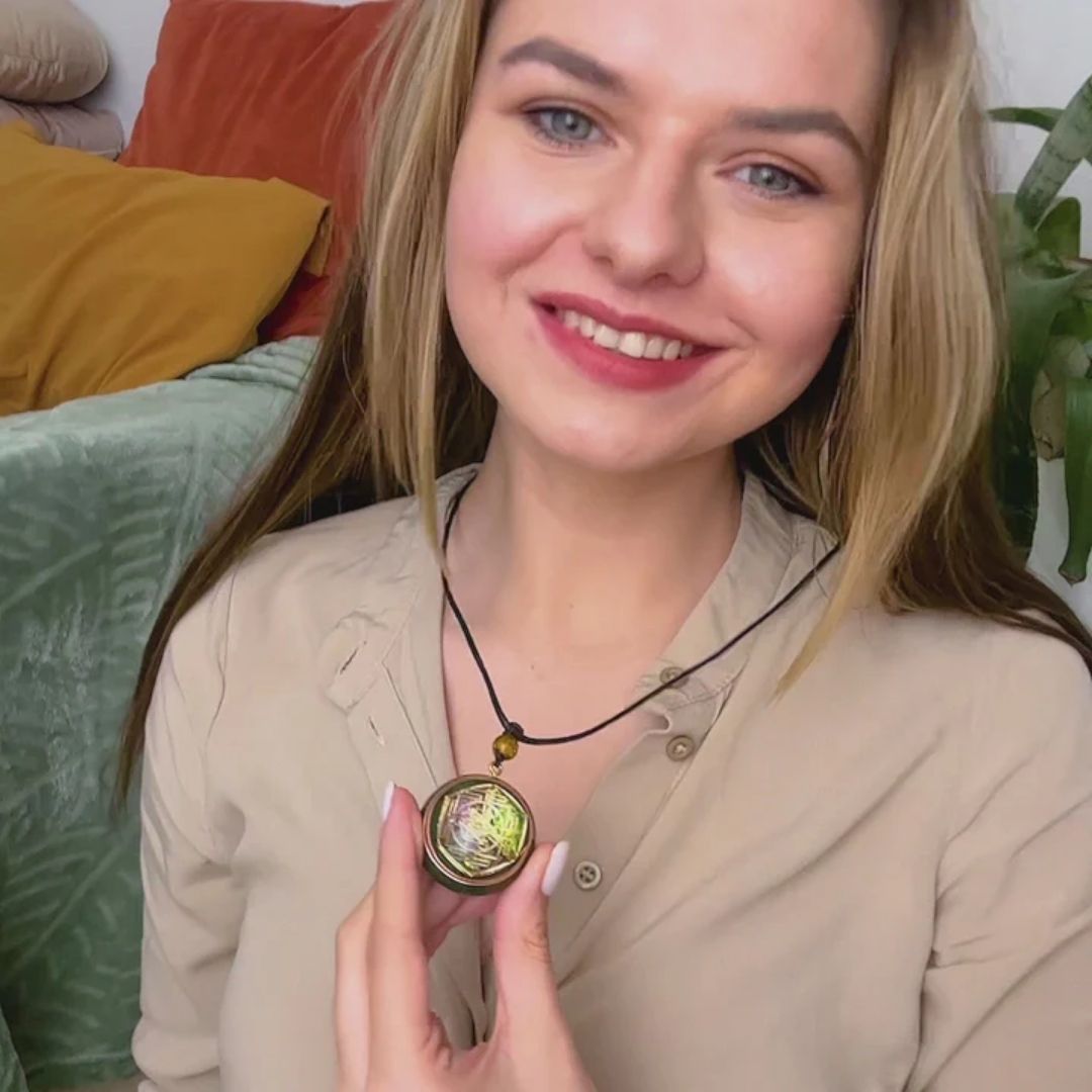 Una mujer sonriente de pelo largo y rubio sostiene el Collar Talismán energético con geometría sagrada, lleva un top beige y está sentada en un sofá con cojines de colores y una planta verde detrás.