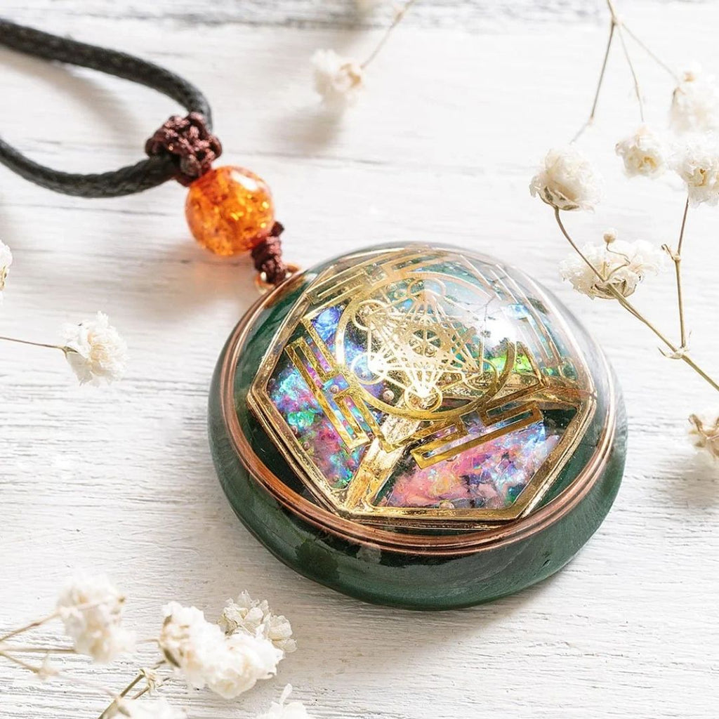 El collar Talismán energético con geometría sagrada, con irisaciones de colores y motivos geométricos dorados, descansa sobre una superficie de madera blanca con delicadas flores blancas. Cuelga de un cordón negro acentuado por una cuenta de ámbar translúcido.