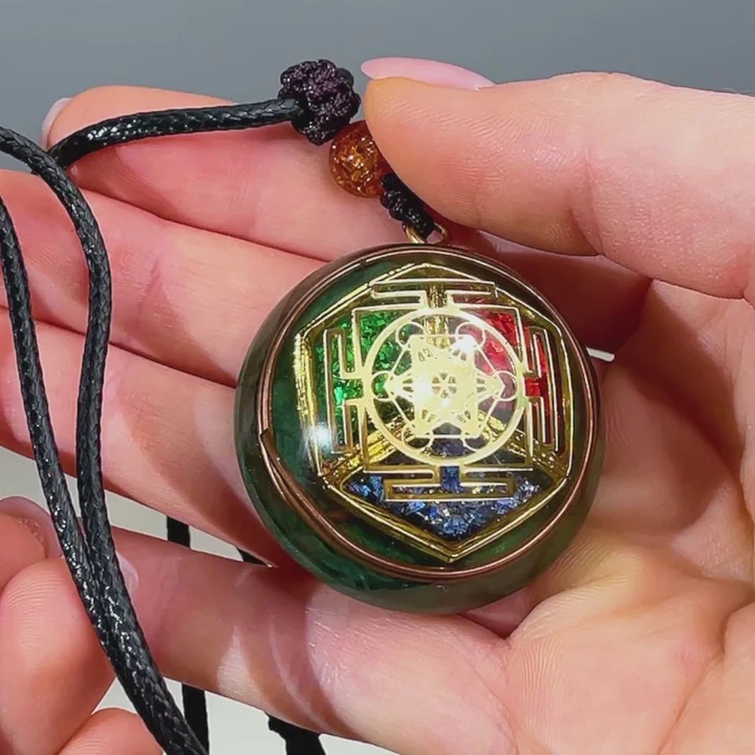 Una mano sujeta el collar Talismán energético con geometría sagrada en un cordón negro, que muestra un diseño geométrico dorado con detalles en rojo, verde y azul y un intrincado dibujo central para el bienestar emocional.