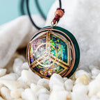 Primer plano del collar Talismán energético con geometría sagrada, un colgante redondo con motivos geométricos y detalles metálicos, con un centro multicolor iridiscente para el bienestar emocional, que cuelga de un cordón negro sobre piedras blancas lisas.
