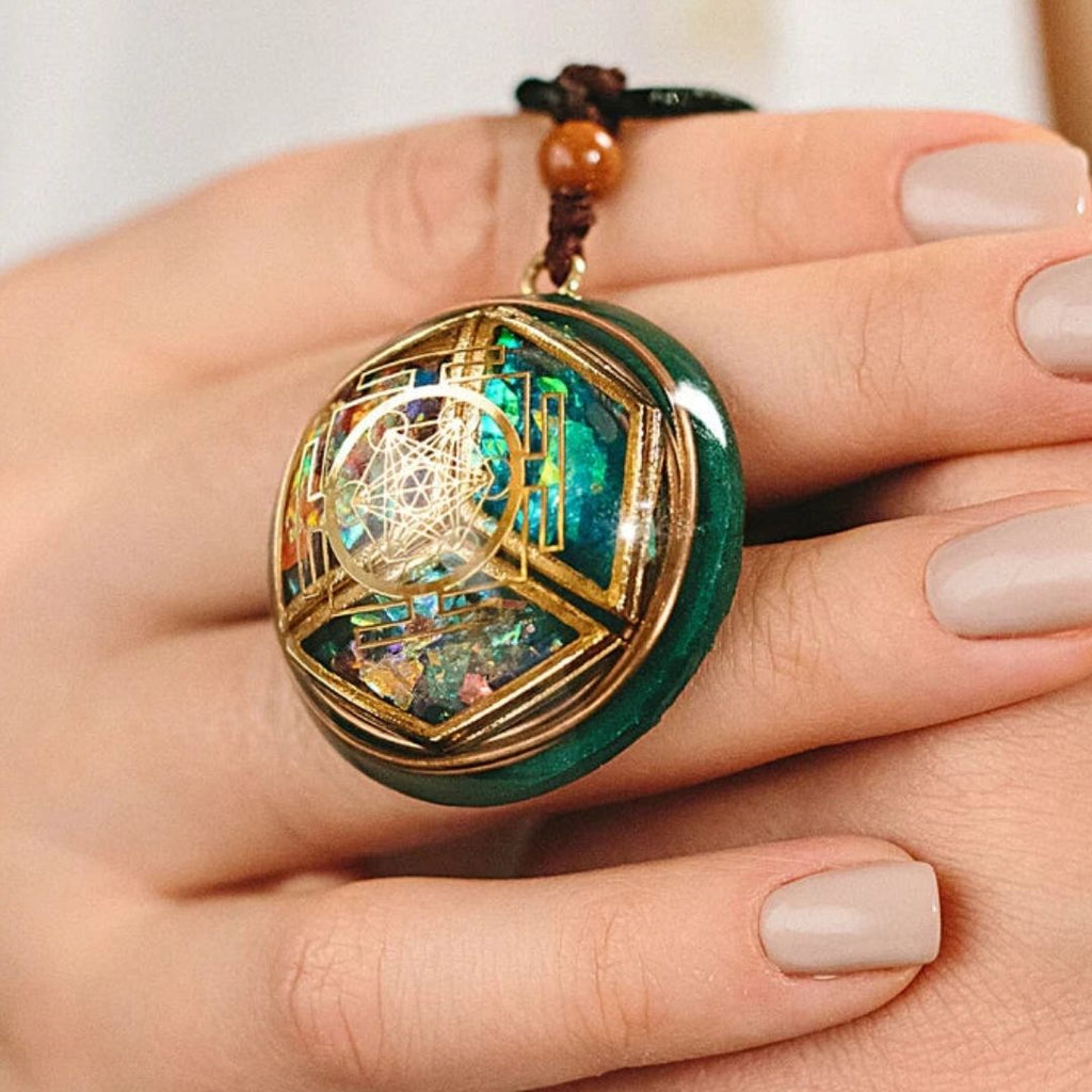 Una mano con las uñas cuidadas sostiene un collar Talismán energético con geometría sagrada, con motivos geométricos dorados y vibrantes detalles turquesa y opalescentes, diseñado para inspirar equilibrio interior y bienestar emocional.