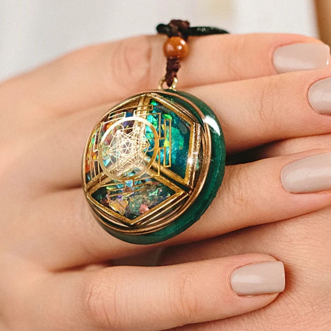 Una mano con las uñas cuidadas sostiene un collar Talismán energético con geometría sagrada, con motivos geométricos dorados y vibrantes detalles turquesa y opalescentes, diseñado para inspirar equilibrio interior y bienestar emocional.