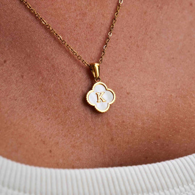 Un primer plano del collar Trébol con inicial - ¡Llévate 2 por el precio de 1!, con un colgante de concha natural en forma de trébol de cuatro hojas con inicial dorada, que se lleva sobre un top blanco de canalé.