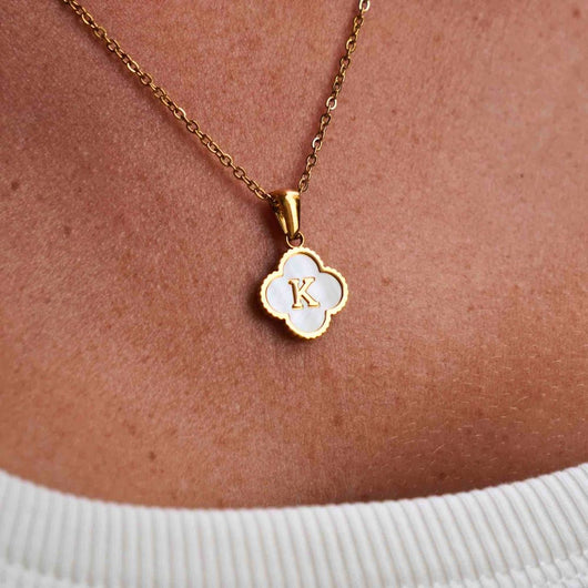 Un primer plano del collar Trébol con inicial - ¡Llévate 2 por el precio de 1!, con un colgante de concha natural en forma de trébol de cuatro hojas con inicial dorada, que se lleva sobre un top blanco de canalé.