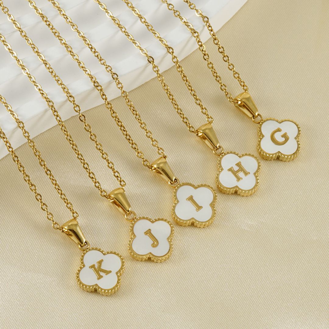 Cinco collares de oro con colgantes de trébol blanco y diferentes letras de oro están dispuestos cerca de una tela blanca-perfecto como un Collar Trébol con inicial. ¡Lévate 2 por el precio de 1!.