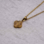 El collar Trébol con inicial - ¡Lévate 2 por el precio de 1! es una cadena de acero inoxidable dorado con un colgante de trébol de cuatro hojas texturizado, que se muestra sobre un fondo de tela gris claro texturizado.