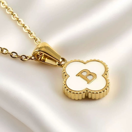 Collar Trébol con inicial - ¡Lévate 2 por el precio de 1! Collar dorado de acero inoxidable con dije de trébol concha natural, incrustación blanca y letra B en el centro, exhibido sobre tela blanca sedosa.