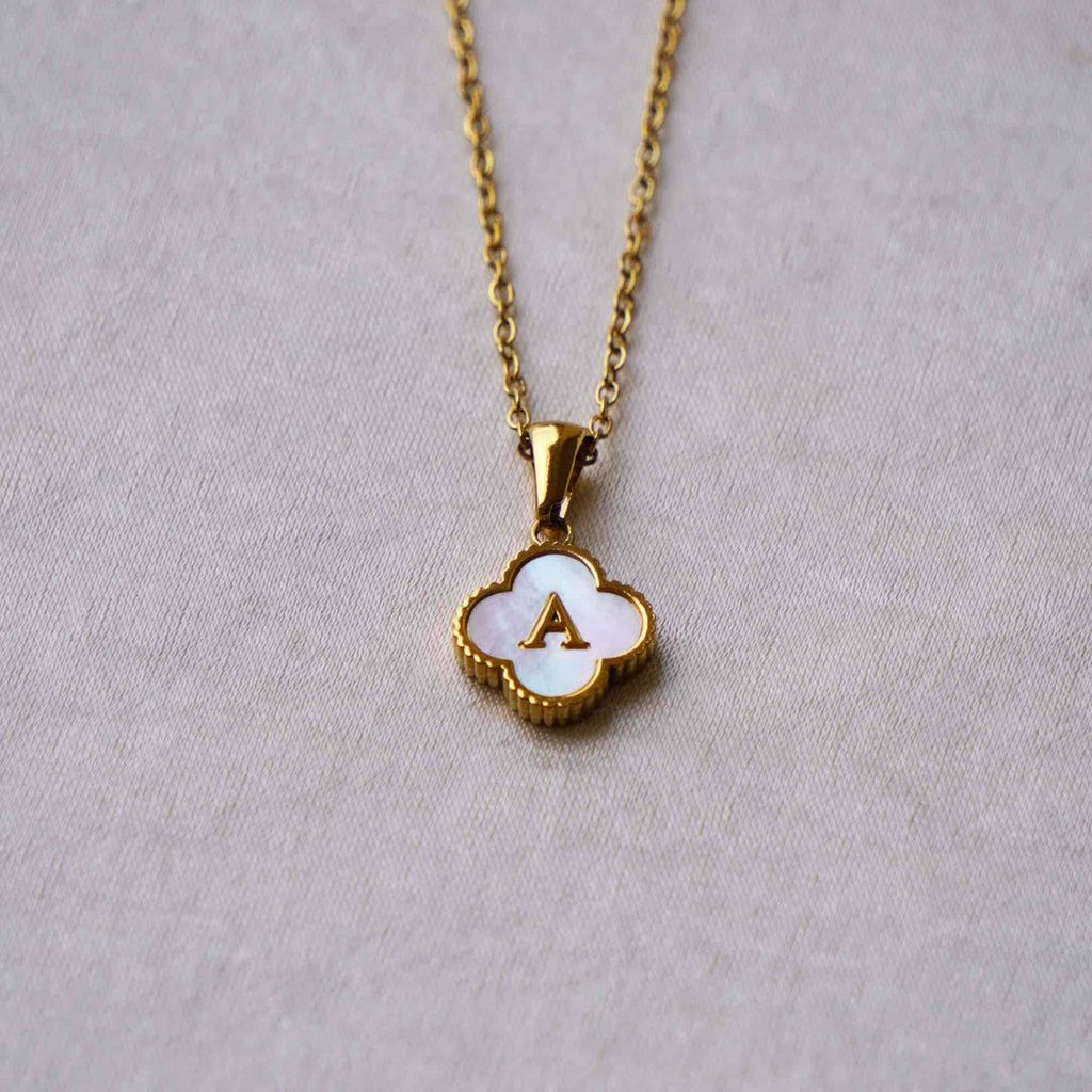 Collar Trébol con inicial: collar de acero inoxidable con dije de concha en forma de trébol, centro blanco y letra A dorada, sobre fondo gris claro. ¡Llévate 2 por el precio de 1!.