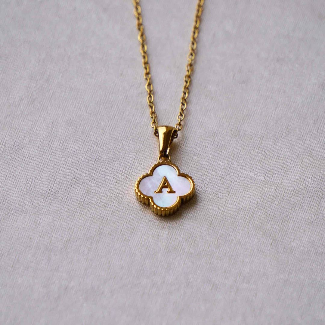 Collar Trébol con inicial: collar de acero inoxidable con dije de concha en forma de trébol, centro blanco y letra A dorada, sobre fondo gris claro. ¡Llévate 2 por el precio de 1!.