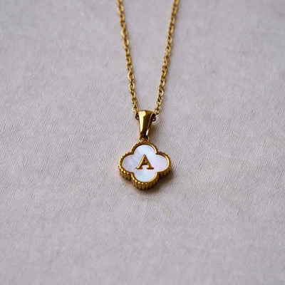 Collar Trébol con inicial: collar de acero inoxidable con dije de concha en forma de trébol, centro blanco y letra A dorada, sobre fondo gris claro. ¡Llévate 2 por el precio de 1!.