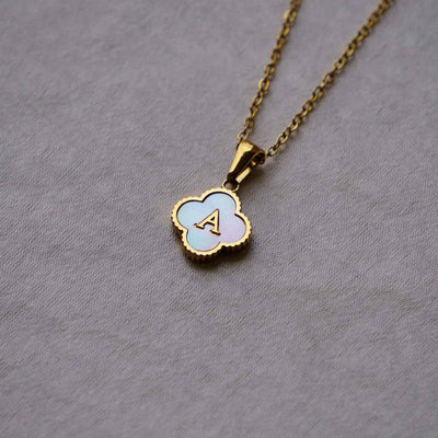 Collar Trébol con inicial - ¡Lévate 2 por el precio de 1! Collar de acero inoxidable con dije de trébol, esmalte blanco y la inicial A dorada, ideal para personalizar tu estilo. Se muestra sobre fondo gris claro y texturizado.