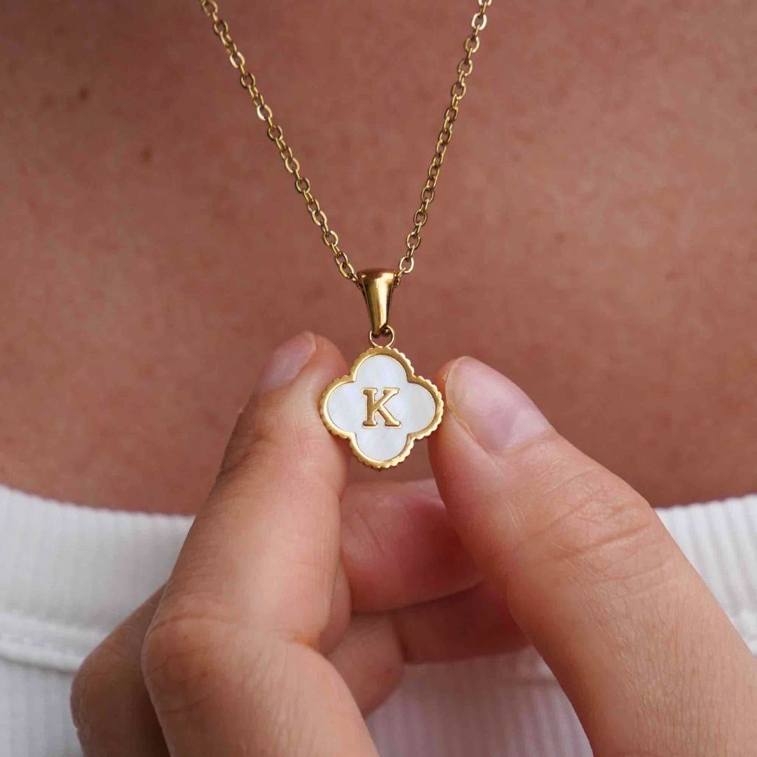 Una persona sostiene cerca de su pecho un Collar Trébol con inicial, que lleva un dije en forma de trébol con la letra K sobre esmalte blanco, mientras usa una blusa blanca. ¡Lévate 2 por el precio de 1!.