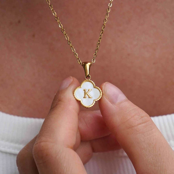 Una persona sostiene cerca de su pecho un Collar Trébol con inicial, que lleva un dije en forma de trébol con la letra K sobre esmalte blanco, mientras usa una blusa blanca. ¡Lévate 2 por el precio de 1!.