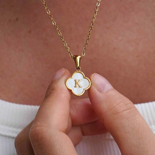 Una persona sostiene cerca de su pecho un Collar Trébol con inicial, que lleva un dije en forma de trébol con la letra K sobre esmalte blanco, mientras usa una blusa blanca. ¡Lévate 2 por el precio de 1!.
