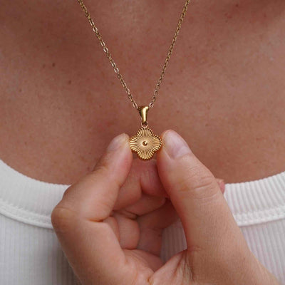 Una persona con blusa blanca sostiene cerca de su pecho el Collar Trébol con inicial - ¡Llévate 2 por el precio de 1!, un collar dorado de acero inoxidable con dije redondo, textura radial y detalle central.