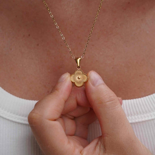 Una persona con blusa blanca sostiene cerca de su pecho el Collar Trébol con inicial - ¡Llévate 2 por el precio de 1!, un collar dorado de acero inoxidable con dije redondo, textura radial y detalle central.