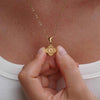 Una persona con blusa blanca sostiene cerca de su pecho el Collar Trébol con inicial - ¡Llévate 2 por el precio de 1!, un collar dorado de acero inoxidable con dije redondo, textura radial y detalle central.