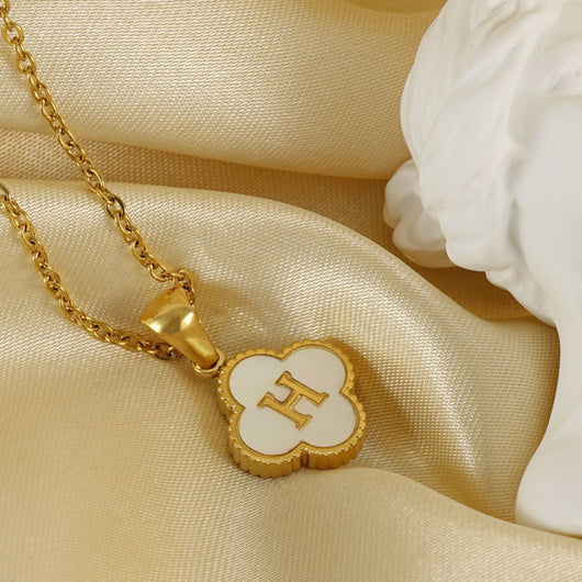 El Collar Trébol con inicial - ¡Llévate 2 por el precio de 1! luce una letra personalizada en acero inoxidable y un dije de concha natural sobre satén crema, acompañado de un adorno blanco.