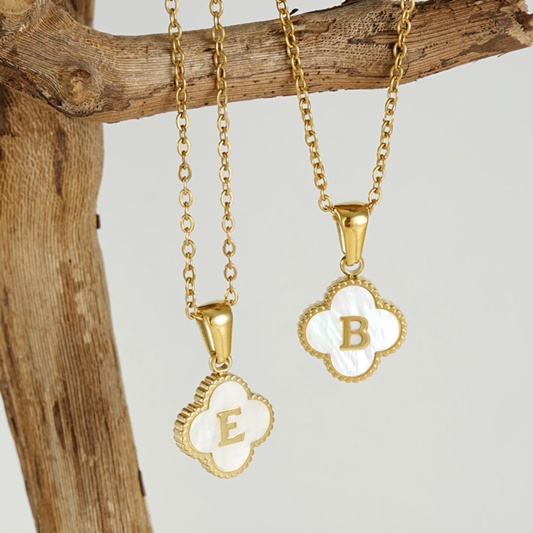 Dos collares Trébol con inicial cuelgan de una rama, cada uno con un charm de concha natural en forma de trébol con las letras "E" y "B" doradas. El fondo neutro resalta estas elegantes piezas. ¡Lévate 2 por el precio de 1!.