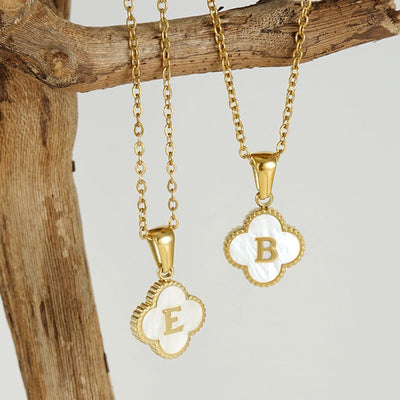Dos collares Trébol con inicial cuelgan de una rama, cada uno con un charm de concha natural en forma de trébol con las letras "E" y "B" doradas. El fondo neutro resalta estas elegantes piezas. ¡Lévate 2 por el precio de 1!.