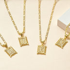 Cuatro collares de oro Marco con inicial personalizada (Oferta 2x1), cada uno con una inicial cuadrada ornamentada colgante-A, J, K, o M-se muestran en una superficie de color beige claro con un objeto rectangular blanco en la esquina.
