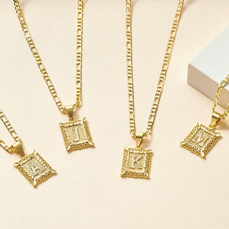 Cuatro collares de oro Marco con inicial personalizada (Oferta 2x1), cada uno con una inicial cuadrada ornamentada colgante-A, J, K, o M-se muestran en una superficie de color beige claro con un objeto rectangular blanco en la esquina.