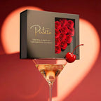 Una copa de martini con una cereza se balancea sobre la caja negra de Experiencia Perlitta - Ostra real, adornada con rosas rojas y letras doradas, evocando la emoción de las ostras con perla, un verdadero regalo experiencial. Una gran forma de corazón se difumina en el fondo rojo.