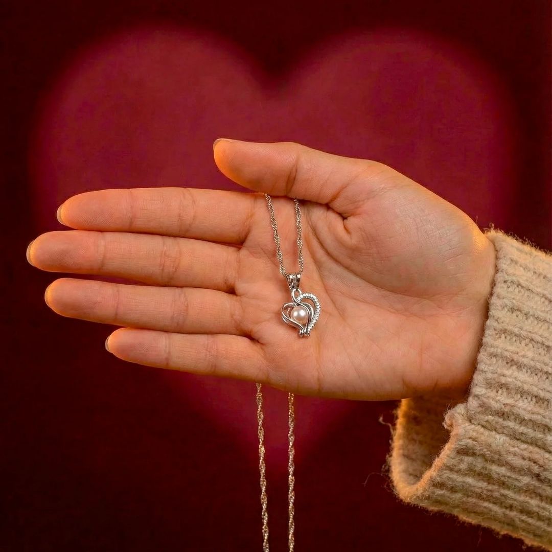 Una mano enfundada en un jersey beige sostiene la Experiencia Perlitta - Ostra real: un collar colgante de plata en forma de corazón, eco de ostras con perla, sobre un fondo rojo oscuro con un corazón resplandeciente: un regalo experiencial ideal.