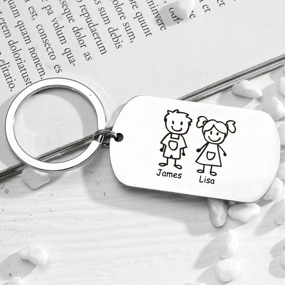 El Llavero Familia personalizable es un llavero metálico con una etiqueta rectangular en la que aparecen unos sencillos dibujos lineales de un niño llamado James y una niña llamada Lisa, expuestos sobre una superficie de madera blanca con piedras blancas esparcidas.