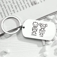El Llavero Familia personalizable es un llavero metálico con una etiqueta rectangular en la que aparecen unos sencillos dibujos lineales de un niño llamado James y una niña llamada Lisa, expuestos sobre una superficie de madera blanca con piedras blancas esparcidas.