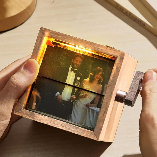 Una persona sostiene un Marco fotográfico con manivela, que muestra una foto iluminada de una pareja sonriente en su boda al aire libre, convirtiéndolo en un regalo conmemorativo significativo.