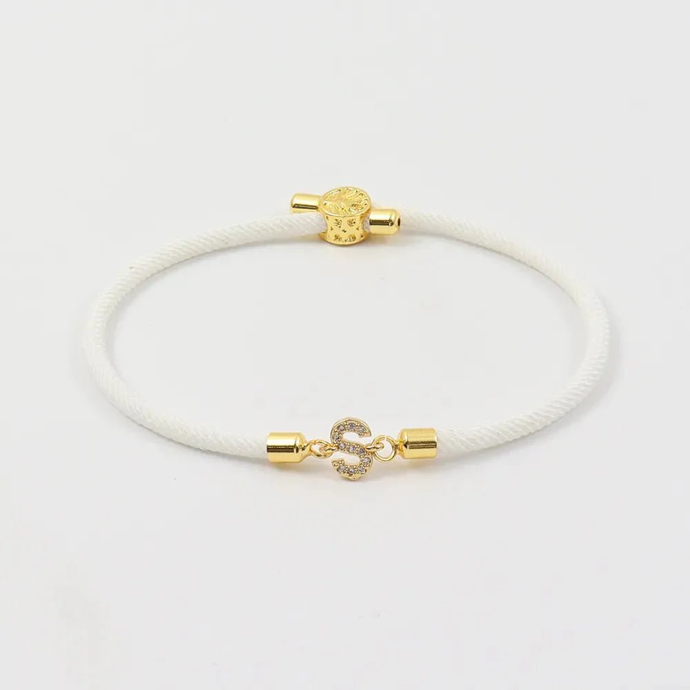 A Pulsera ajustable con inicial (2X1) en color blanco, con extremos metálicos dorados y broche en forma de S decorado con gemas brillantes, exhibida sobre fondo blanco.