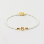 A Pulsera ajustable con inicial (2X1) en color blanco, con extremos metálicos dorados y broche en forma de S decorado con gemas brillantes, exhibida sobre fondo blanco.