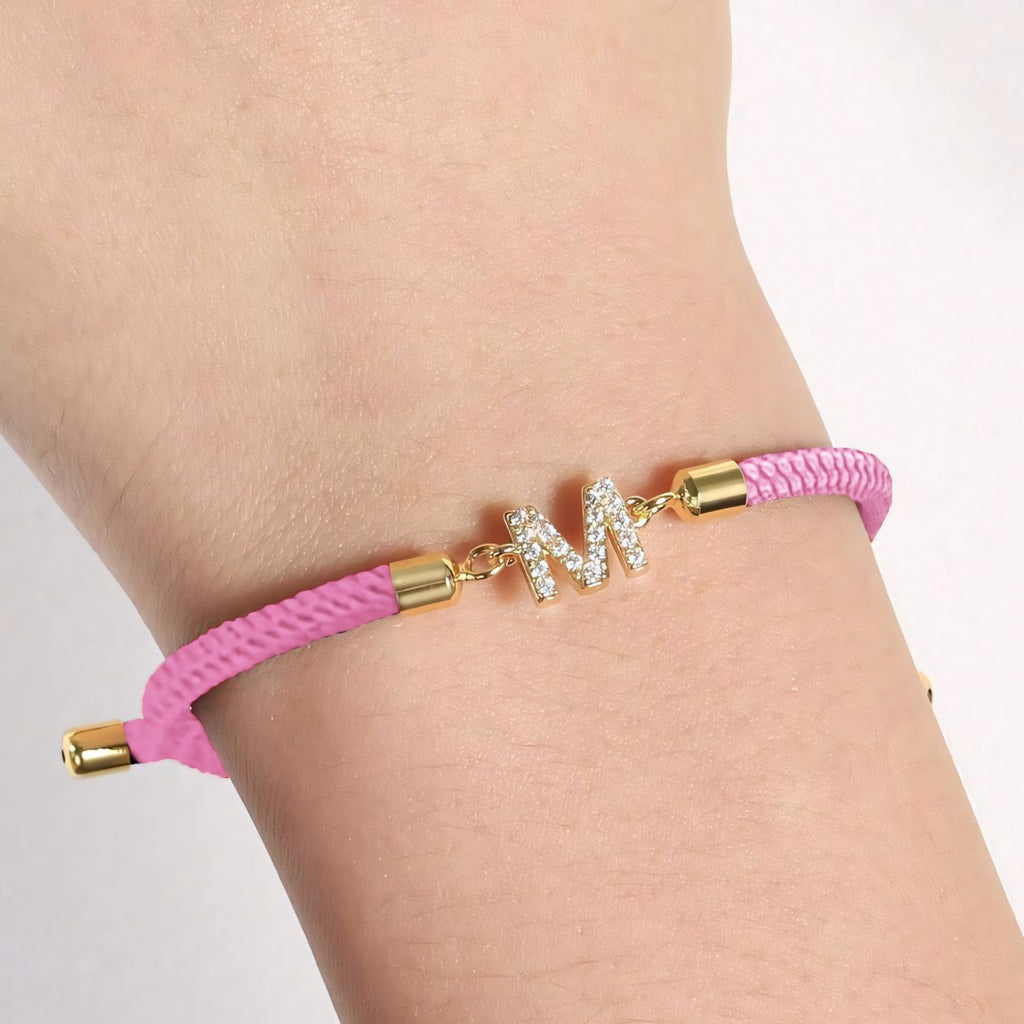 Luce tu estilo único con la Pulsera ajustable con inicial (2X1): banda rosa trenzada, broches dorados y dije M con brillantes. Aprovecha la oferta 2x1 y personaliza tu look con esta pulsera exclusiva.