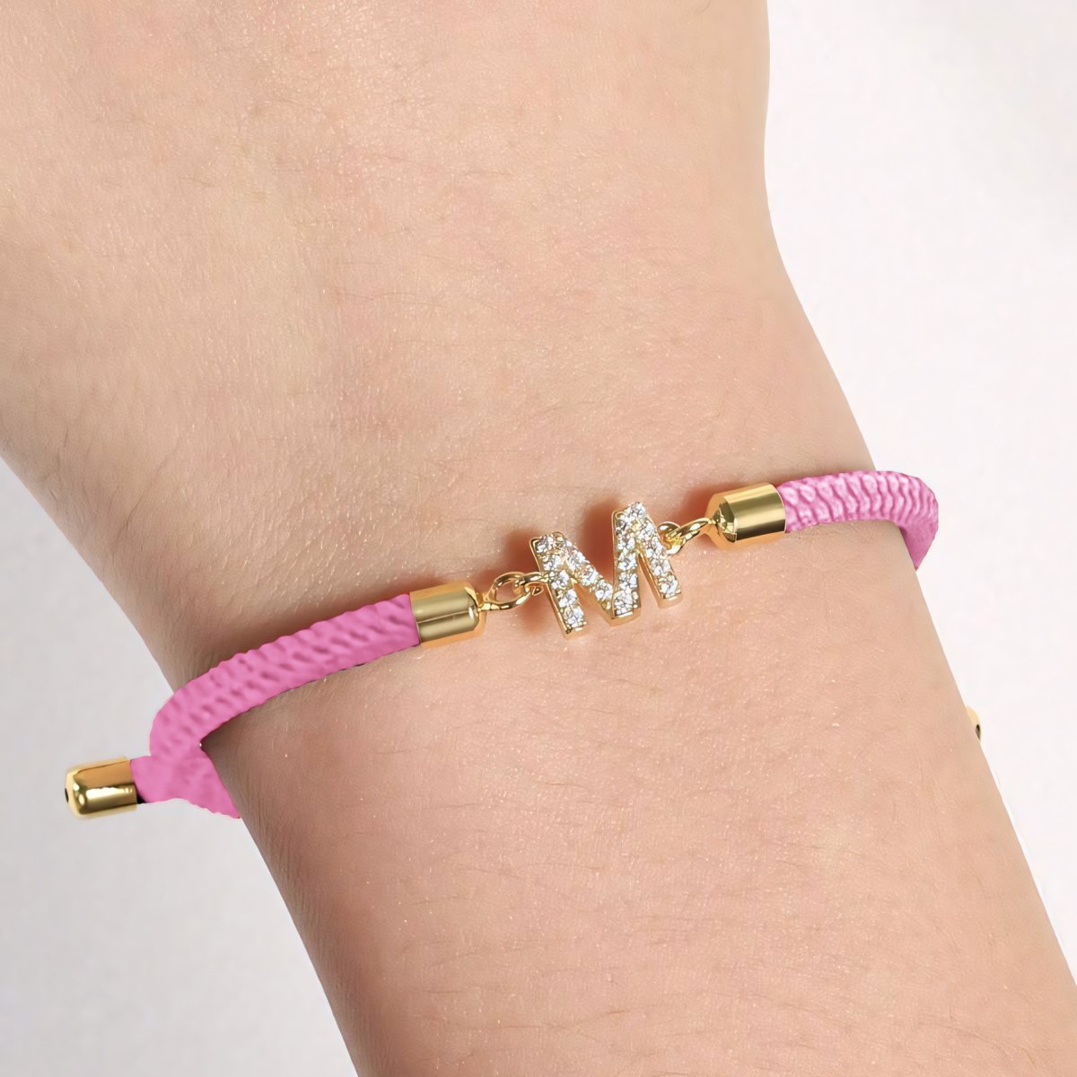 Luce tu estilo único con la Pulsera ajustable con inicial (2X1): banda rosa trenzada, broches dorados y dije M con brillantes. Aprovecha la oferta 2x1 y personaliza tu look con esta pulsera exclusiva.