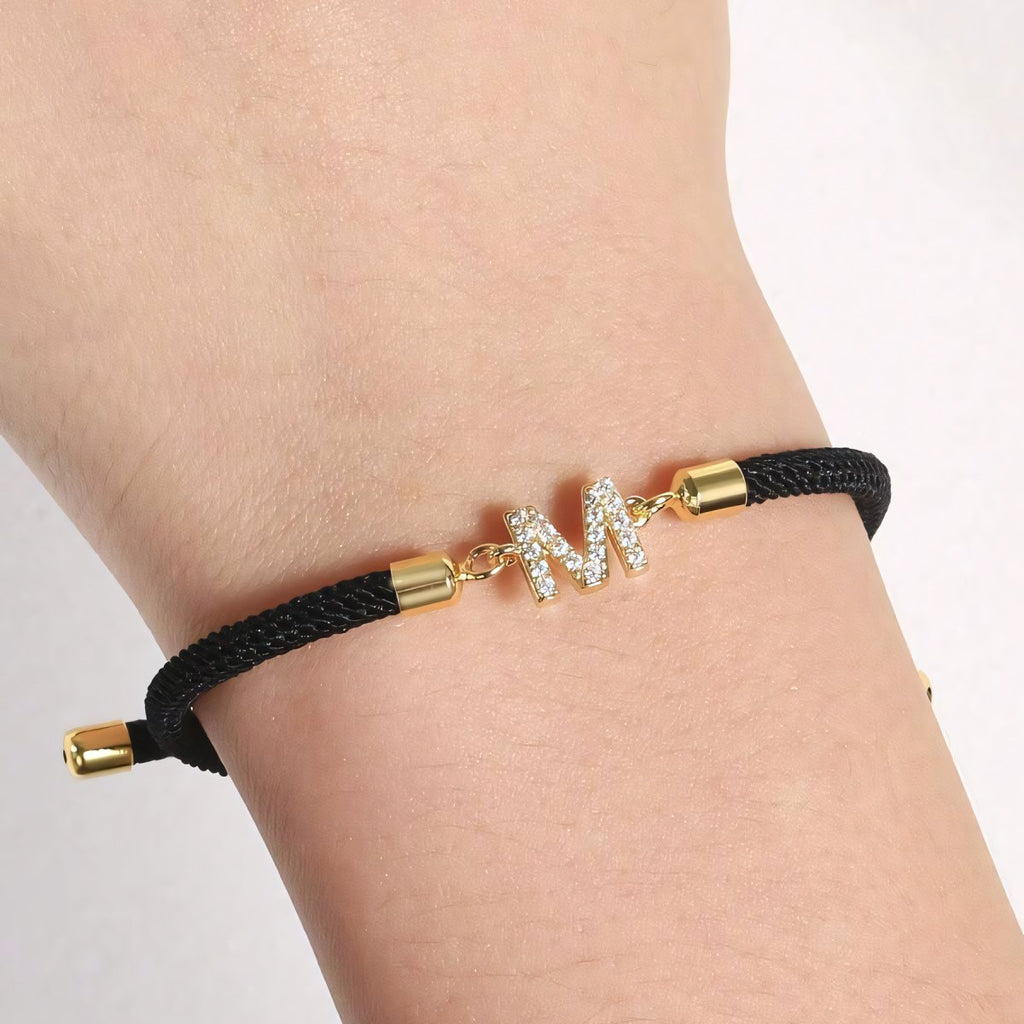 Luce tu Pulsera ajustable con inicial (2X1): diseño con cordón negro, terminales doradas y dije en forma de M decorado con piedras claras. ¡Aprovecha la oferta 2x1 en tu pulsera personalizada!.