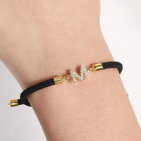 Luce tu Pulsera ajustable con inicial (2X1): diseño con cordón negro, terminales doradas y dije en forma de M decorado con piedras claras. ¡Aprovecha la oferta 2x1 en tu pulsera personalizada!.