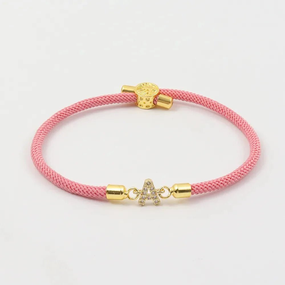 Pulsera ajustable con inicial (2X1) color rosa, trenzada y realzada con detalles dorados y charm de letra A en cristal. ¡Aprovecha la oferta 2x1 y lleva tu pulsera personalizada!.