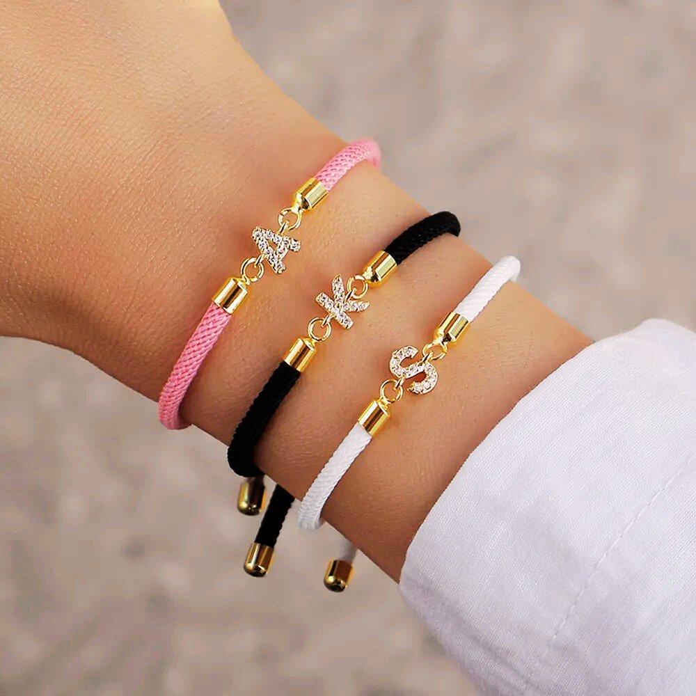 Una mano luce tres Pulseras ajustables con inicial (2X1): rosa con "A", negra con "K" y blanca con "S", todas con detalles dorados y brillantes. ¡Aprovecha la oferta 2x1 en tu pulsera favorita!.