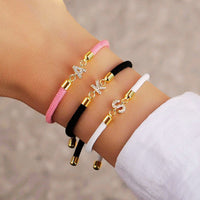 Una mano luce tres Pulseras ajustables con inicial (2X1): rosa con "A", negra con "K" y blanca con "S", todas con detalles dorados y brillantes. ¡Aprovecha la oferta 2x1 en tu pulsera favorita!.
