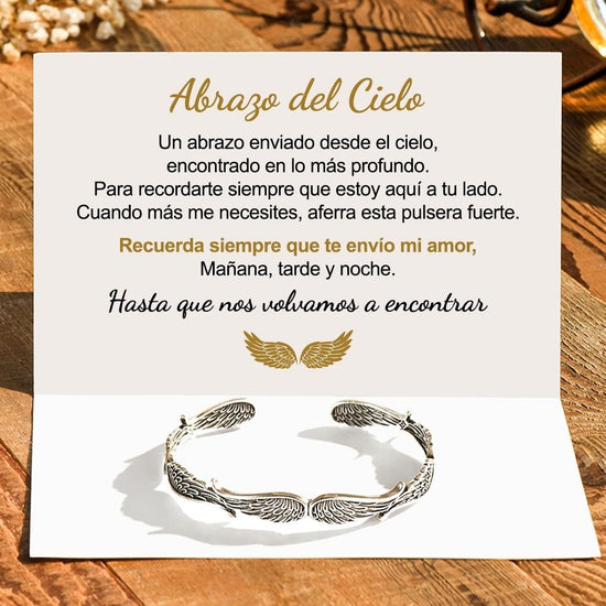 Una tarjeta con un mensaje en español sobre un abrazo del cielo se abre sobre madera, acompañada de la Pulsera Alas de ángel -símbolo de protección. La tarjeta muestra alas doradas y palabras reconfortantes, lo que la convierte en un regalo Abrazo del Cielo ideal.