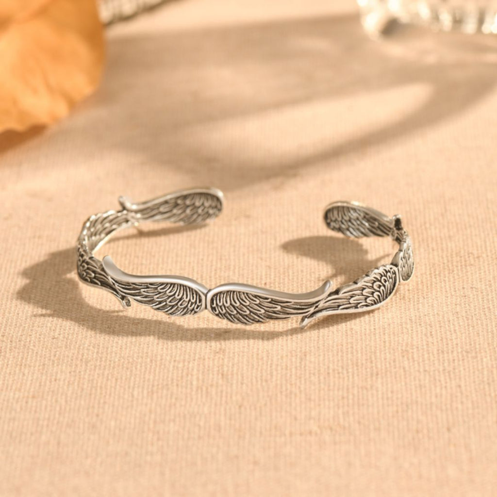 La Pulsera Alas de ángel es una pulsera de plata con detallados diseños de alas emplumadas, mostrada sobre tela beige con suave luz natural y fondo difuminado.