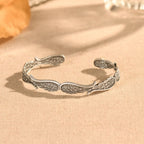 La Pulsera Alas de ángel es una pulsera de plata con detallados diseños de alas emplumadas, mostrada sobre tela beige con suave luz natural y fondo difuminado.