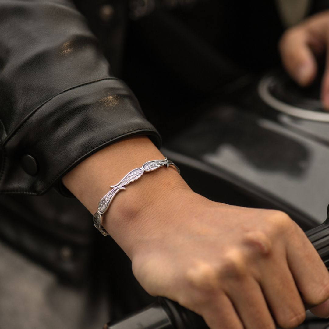 Primer plano de una persona que lleva la Pulsera Alas de ángel, un brazalete de plata y una chaqueta de cuero negro mientras se agarra al manillar de una motocicleta negra.